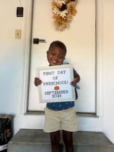 A young boy proudly holds a sign that reads "First Day of Preschool," smiling with excitement for his new adventure.