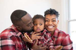 A joyful black couple stands with their child in a red plaid shirt, enjoying a festive atmosphere together.
